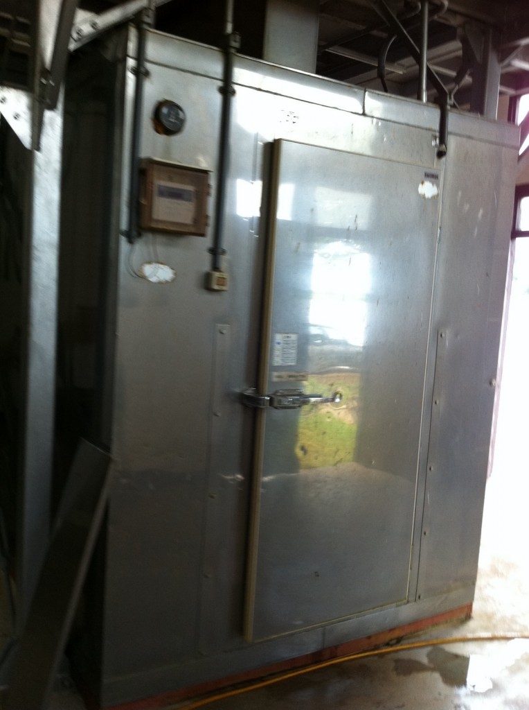 The image shows a large, industrial metal cabinet or enclosure. It has a door with a handle and a latch, and there are various pipes and electrical conduits attached to it. There is a gauge and a small control panel mounted on the front. The enclosure appears to be part of a larger industrial setup, possibly used for processes such as heating, cooling, or storage. The surrounding area looks like an industrial or manufacturing facility.