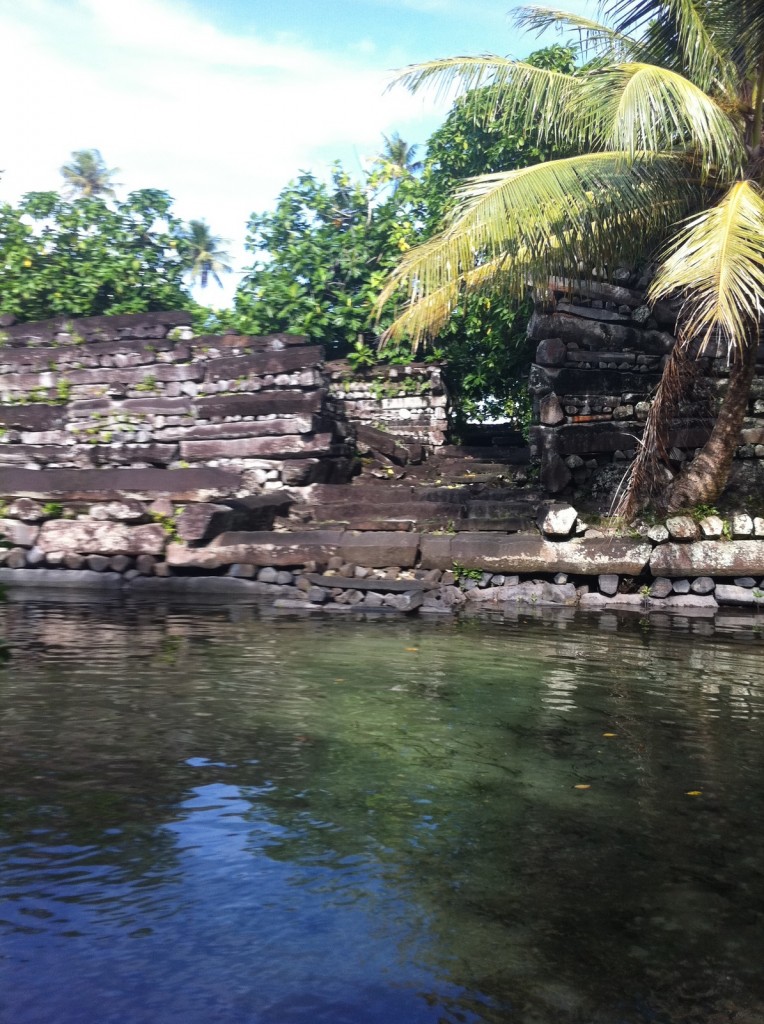 The image shows an ancient stone structure partially submerged in clear water. The structure is made of stacked stones and appears to be in a state of ruin. Surrounding the structure are lush green trees, including a prominent palm tree on the right side. The sky above is clear with a few clouds, indicating a sunny day. The water in the foreground is calm and reflects the surrounding greenery and stone structure.