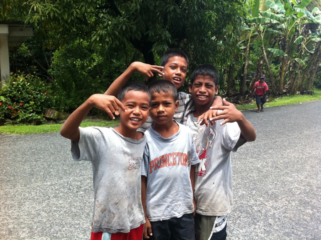 Four boys are standing on a paved road, smiling and posing for the camera. They are wearing casual clothes, with one boy wearing a shirt that says "Princeton." Behind them, there is lush greenery with trees and plants, and another person can be seen walking in the background. The scene appears to be in a rural or suburban area.