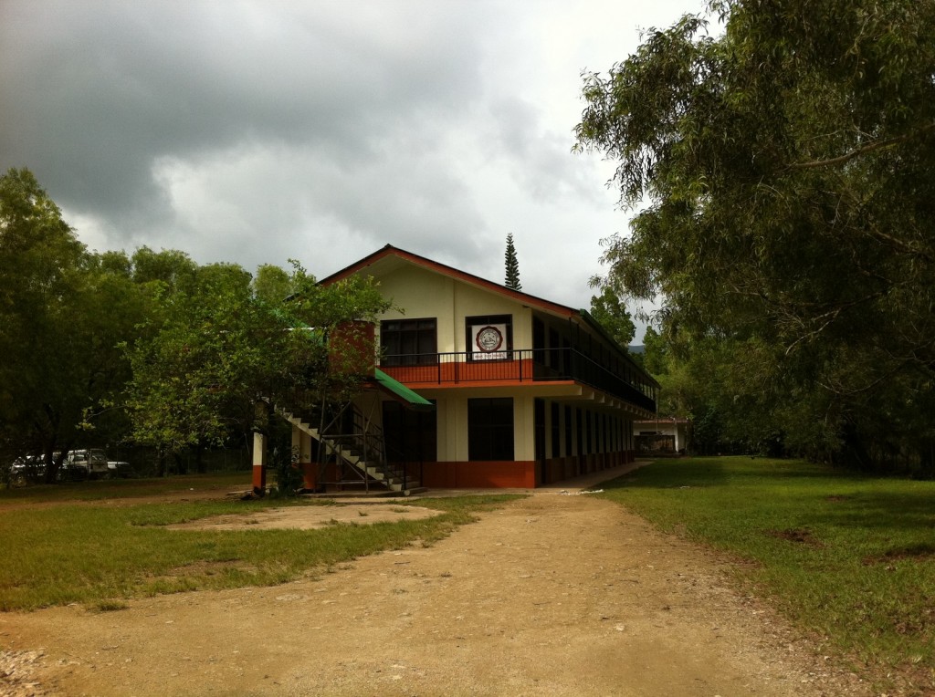 A two-story building with a red roof and white walls, featuring a balcony on the upper floor. The building is surrounded by trees and greenery, with a dirt path leading up to it. The sky is overcast with dark clouds. There is a sign on the upper floor of the building, but the text is not clearly visible.