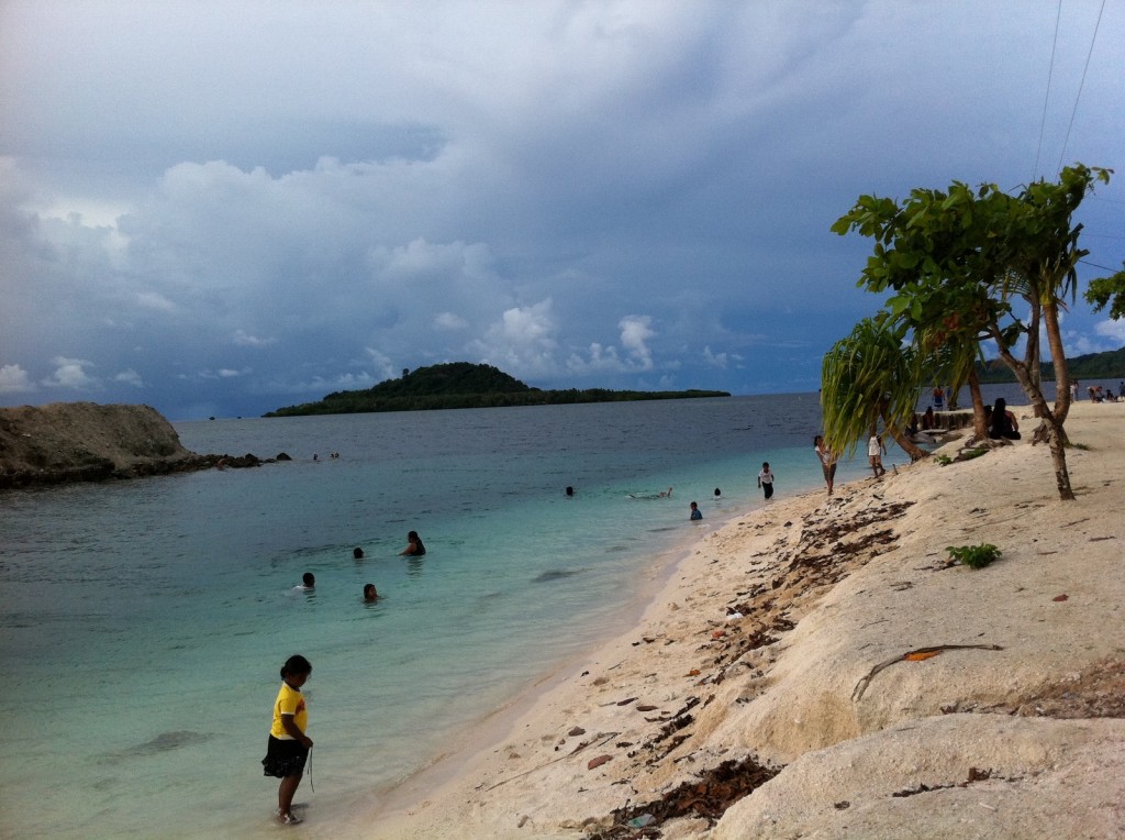 The image shows a beach scene with several people enjoying the water and the shore. The beach has light-colored sand and is lined with some trees and vegetation. The water is clear and transitions from light to dark blue as it gets deeper. In the background, there is a small island covered with greenery. The sky is mostly cloudy, suggesting an overcast day. Some people are swimming in the water, while others are standing or walking along the beach.