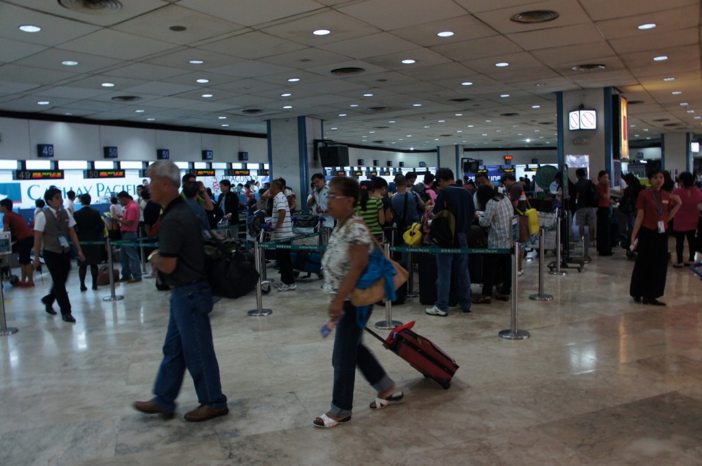 The image shows a busy airport check-in area with many people standing in line at various counters. The counters are numbered and have signs above them, with some displaying the name "Cathay Pacific." People are seen with luggage, some pulling suitcases, and others carrying bags. The floor is made of light-colored tiles, and the ceiling has numerous recessed lights. There are also stanchions with belts to organize the lines. The atmosphere appears bustling, typical of an airport setting.