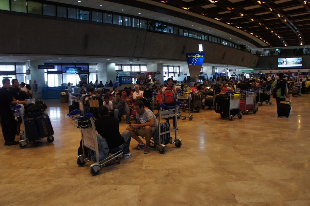 The image shows a busy airport terminal with many people sitting and standing around. Some people are seated on chairs, while others are sitting on luggage carts. There are numerous luggage carts filled with suitcases and bags. The terminal has a high ceiling with many lights, and there is an information sign in the center of the image. In the background, there are various counters and signs, including one for "Travel Tax." The overall atmosphere is bustling, with travelers waiting and moving around.