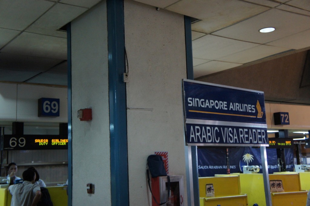 The image shows a section of an airport terminal. There is a sign for "Singapore Airlines" with "Arabic Visa Reader" written below it. In the background, there are gate numbers 69 and 72 visible. There is also a digital display with the text "SAUDIA DUTY AIRLINES OFFICER." Some people are seen in the background near the counters. The overall setting appears to be a check-in or boarding area.