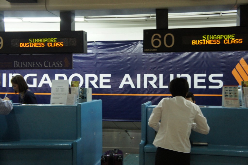The image shows a check-in counter for Singapore Airlines at an airport. There are two counters labeled "Business Class" with digital signs displaying "Singapore Business Class." Two people are being assisted by airline staff at the counters. The background features a large banner with the Singapore Airlines logo and name.