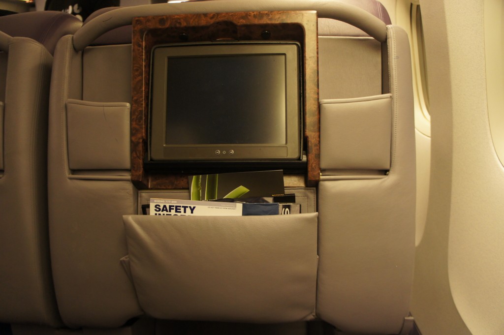 The image shows the back of an airplane seat. There is an in-flight entertainment screen mounted on the back of the seat, surrounded by a wooden frame. Below the screen, there is a pocket containing a safety instruction card and other informational materials. The seat is upholstered in gray material, and part of the airplane window is visible on the right side of the image.
