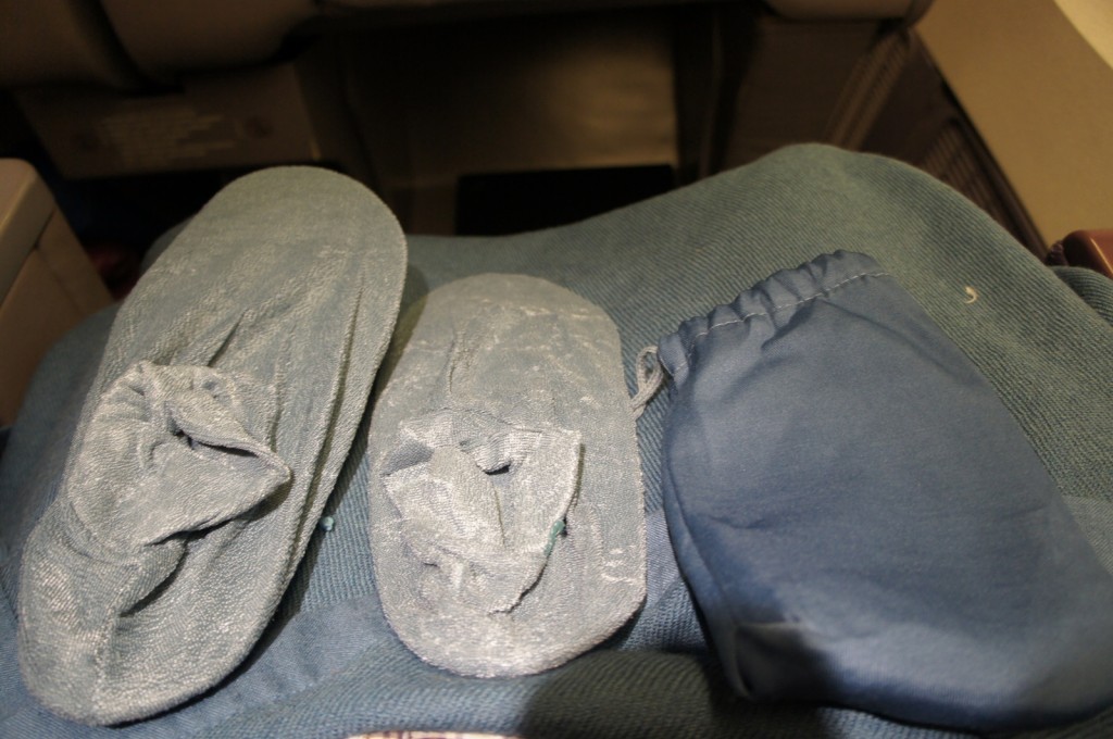 The image shows a pair of gray disposable slippers and a small blue drawstring bag placed on a blue fabric surface, likely on an airplane seat.