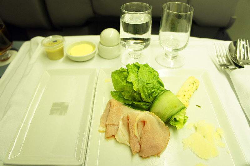 The image shows a meal served on an airplane tray table. The meal consists of slices of ham, lettuce, a cucumber slice, and some cheese on a white rectangular plate. There are two glasses, one filled with water and the other empty, placed behind the plate. Additionally, there are small containers of butter and mustard, a salt and pepper shaker, and a set of cutlery wrapped in a napkin on the right side of the tray. The background appears to be the seat and tray table of an airplane.