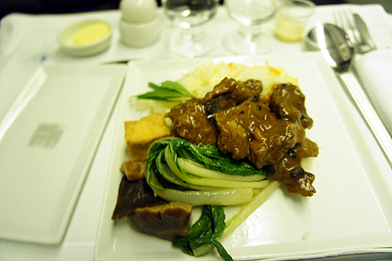 The image shows a plated meal consisting of a meat dish with a brown sauce, served alongside green vegetables, possibly bok choy, and some tofu pieces. There is also a portion of rice on the plate. The meal is presented on a white square plate, and the setting includes a fork, knife, and spoon, along with a glass of water and a small dish of butter in the background. The overall presentation suggests a formal dining setting, possibly on an airplane or in a restaurant.