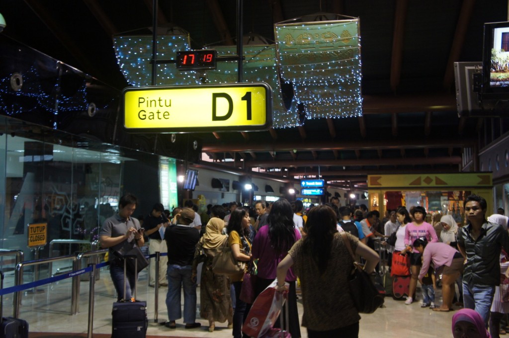 The image shows a busy airport terminal with a crowd of people. A prominent yellow sign overhead indicates "Pintu Gate D1." There is a digital clock above the sign displaying the time as 17:19. The terminal is decorated with string lights, and there are various signs and advertisements visible in the background. People are standing with their luggage, some are checking their phones, and others are engaged in conversation. A "Tutup Closed" sign is visible on the left side of the image.