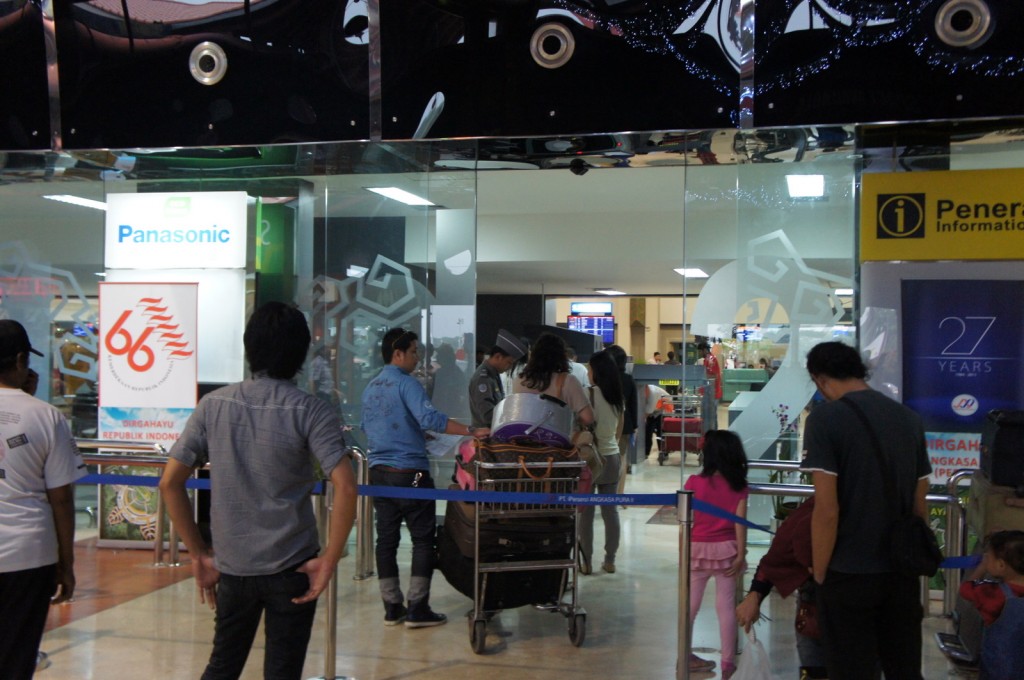 The image shows a group of people standing in line at an airport check-in or information counter. There are signs for "Panasonic" and "Penerangan Information" visible, along with a banner celebrating "66" years. Some people are pushing luggage carts, and there are children among the group. The area is well-lit, and there are glass doors and partitions in the background.