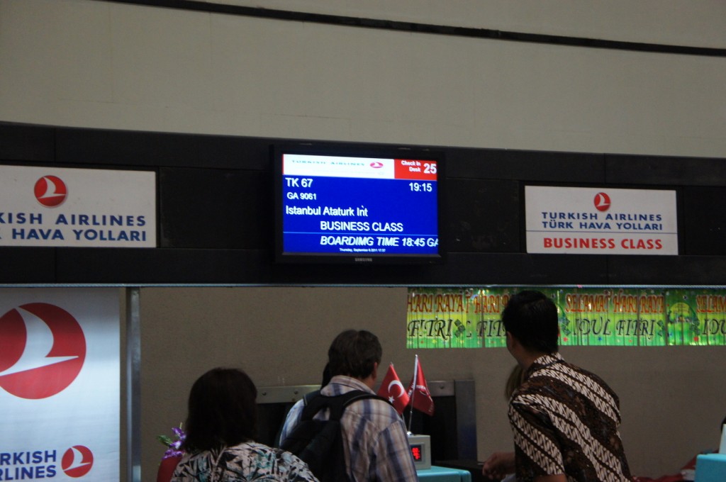 The image shows a Turkish Airlines boarding gate area. There is a digital display screen showing flight information for flight TK 67 to Istanbul Ataturk International Airport. The flight is for business class passengers, with a boarding time of 18:45 and a departure time of 19:15. The check-in desk is number 25. There are signs on either side of the display screen indicating "Turkish Airlines" and "Business Class" in both English and Turkish. Several people are standing in front of the counter, and there are small Turkish flags on the counter.