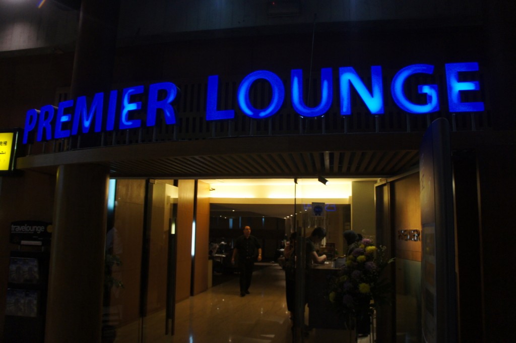 The image shows the entrance to a "Premier Lounge" with a bright blue sign above the doorway. The interior is warmly lit, and there are people inside, some standing at a counter. There is a flower arrangement near the entrance, and a sign on the left side that reads "travelounge." The overall ambiance suggests a comfortable and upscale waiting area, likely in an airport or similar setting.