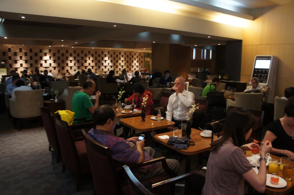 A busy airport lounge with several people sitting at tables and chairs. Some individuals are eating, drinking, or using their phones. The lounge has a modern design with warm lighting and a patterned wall in the background. There are various drinks and food items on the tables, and a self-service kiosk is visible in the background. The atmosphere appears to be relaxed and social.