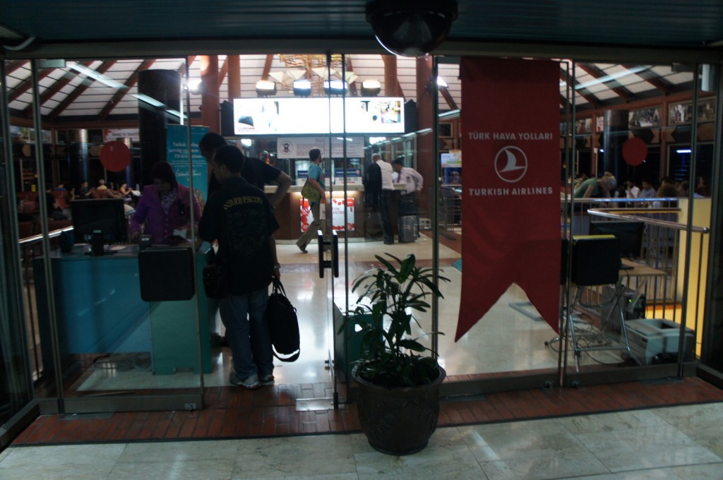 The image shows the entrance to a Turkish Airlines lounge or office area at an airport. The entrance has glass doors, and a red banner with the Turkish Airlines logo and the text "TÜRK HAVA YOLLARI" and "TURKISH AIRLINES" is displayed on the right side. Inside, there are people interacting with staff at a counter. The area is well-lit, and there is a potted plant in front of the entrance. In the background, there are more people and some digital screens displaying information.