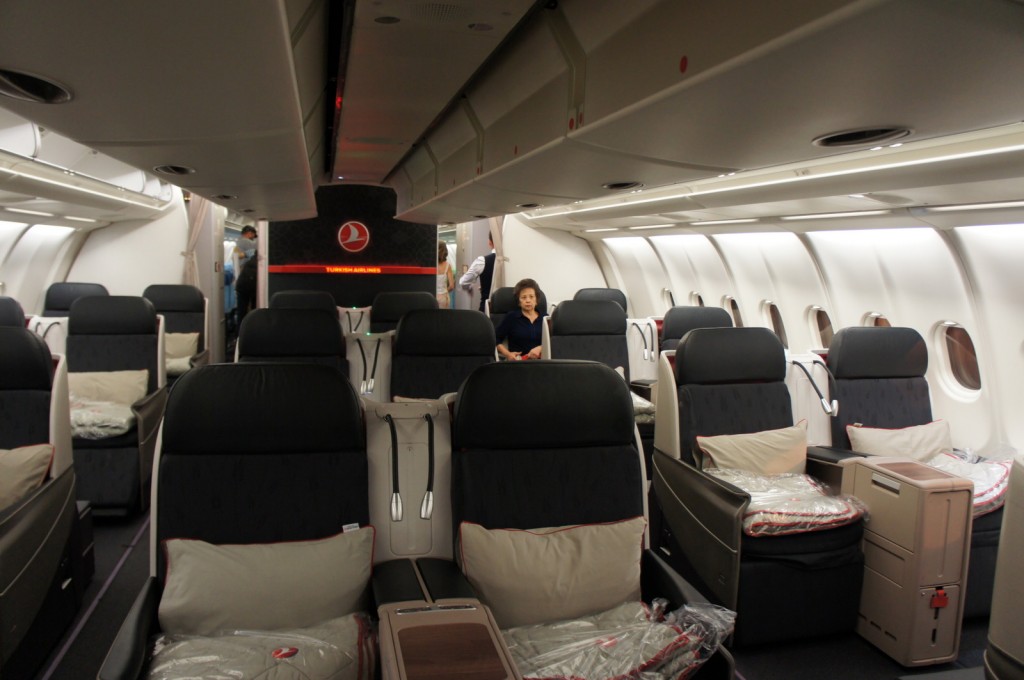 The image shows the interior of a business class cabin on a Turkish Airlines aircraft. The cabin features spacious, comfortable seats arranged in a 2-2-2 configuration. Each seat is equipped with a pillow and a blanket. The cabin is well-lit with overhead lighting, and there are overhead compartments for storage. A few passengers and flight attendants are visible in the background. The Turkish Airlines logo is prominently displayed on a partition at the front of the cabin.