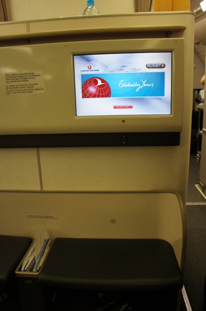 The image shows an in-flight entertainment screen on a Turkish Airlines plane. The screen displays the Turkish Airlines logo and the slogan "Globally Yours" along with the Planet entertainment system logo. Below the screen, there is a small storage area with some documents or magazines. A water bottle is placed on top of the panel above the screen. The surrounding area appears to be part of the aircraft's interior, likely in the business or first-class section.