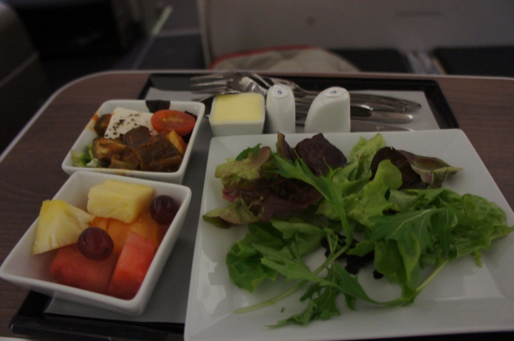 The image shows a meal served on a tray. The meal includes a small bowl of mixed fruit (pineapple, watermelon, and grapes), a small bowl of a dish with pieces of meat, cherry tomatoes, and a piece of cheese, and a plate of mixed green salad. There are also two small containers, likely for salt and pepper, a small square of butter, and a set of cutlery wrapped in a napkin. The tray is placed on a table, possibly in an airplane or a similar setting.