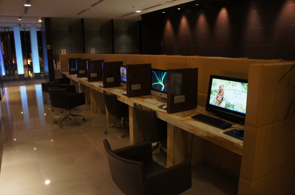 The image shows a modern computer workstation area, likely in a public or business setting such as a hotel or office. There are several individual workstations with desktop computers, each separated by partitions for privacy. Each station has a swivel chair. The room is well-lit with overhead lighting and has a polished floor. The background includes a wall with vertical elements and a section with blue lighting.