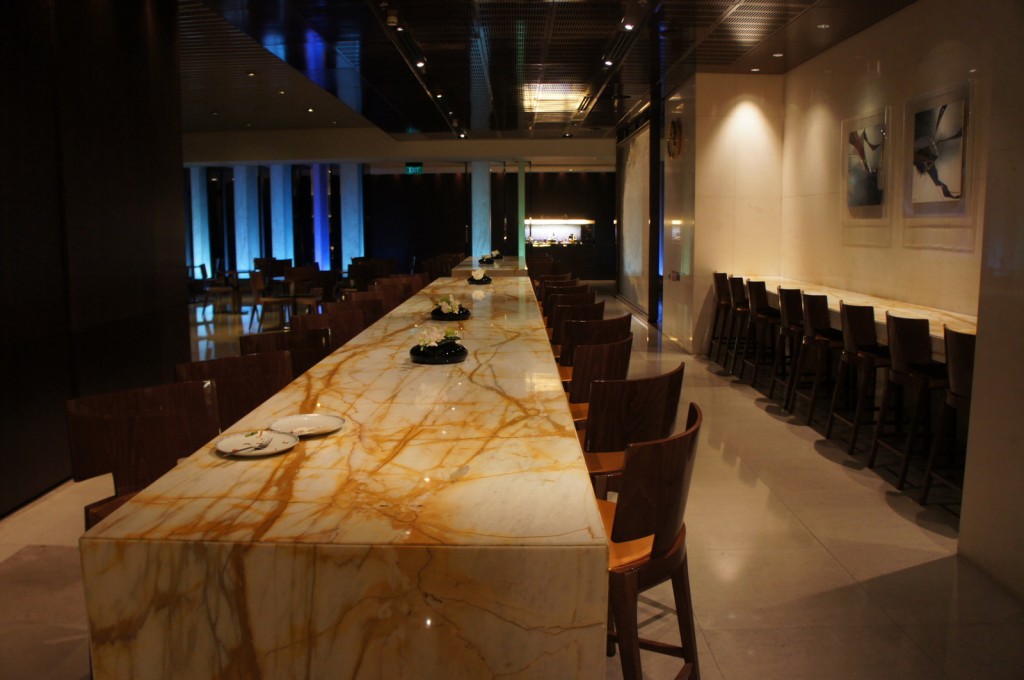 The image shows a modern, upscale dining area with a long, marble-topped communal table in the center. The table is surrounded by wooden chairs, and there are a few small decorative items placed on it. On the right side of the image, there is a long counter with high chairs lined up against it. The walls are adorned with contemporary artwork, and the lighting is dim, creating a cozy and elegant atmosphere. In the background, there are more tables and chairs, and the space appears to be well-lit with a combination of overhead and ambient lighting.
