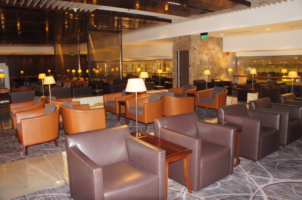 The image shows a spacious and modern lounge area, likely in an airport or hotel. The lounge is furnished with numerous comfortable armchairs in shades of brown and orange, arranged in small groups around wooden tables. There are several floor lamps and table lamps providing warm lighting throughout the space. The floor is carpeted with a patterned design, and the ceiling features a reflective surface. The walls are a mix of glass and stone, and there is an exit sign visible above a door in the background. The overall ambiance is cozy and inviting.