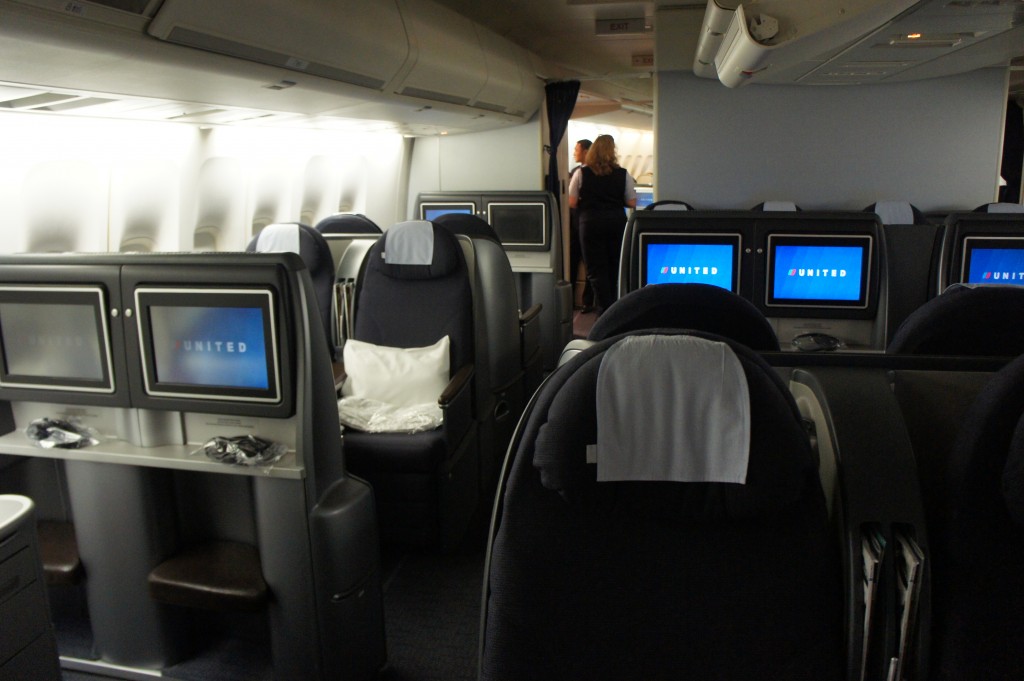 The image shows the interior of a United Airlines business class cabin. The seats are arranged in a 2-2 configuration, with each seat featuring a personal entertainment screen displaying the United logo. The seats are equipped with headrests covered with white cloths, and there are pillows and blankets placed on some of the seats. The cabin has a modern and spacious design, with overhead compartments and a flight attendant visible in the background near the galley area.