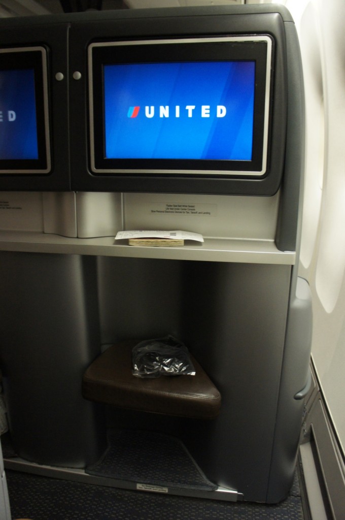 The image shows a seat in a United Airlines aircraft. The seat is equipped with a personal entertainment screen displaying the United Airlines logo. Below the screen, there is a small shelf with a pair of headphones in a plastic bag placed on it. The area around the seat is enclosed, providing a sense of privacy. The overall design is modern and functional, typical of business or first-class seating on an airplane.
