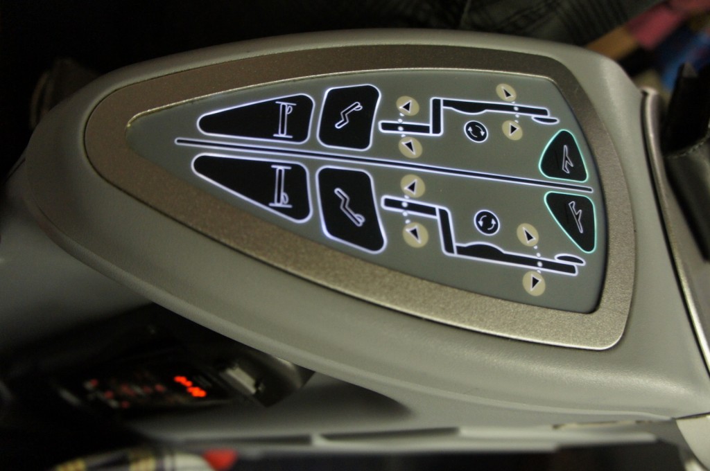 The image shows a close-up of a control panel, likely from an airplane seat. The panel has various buttons with icons indicating different seat adjustments, such as reclining, leg rest extension, and possibly lumbar support. The buttons are backlit, making the icons visible in low light. The panel is embedded in a gray surface, possibly the armrest of the seat.