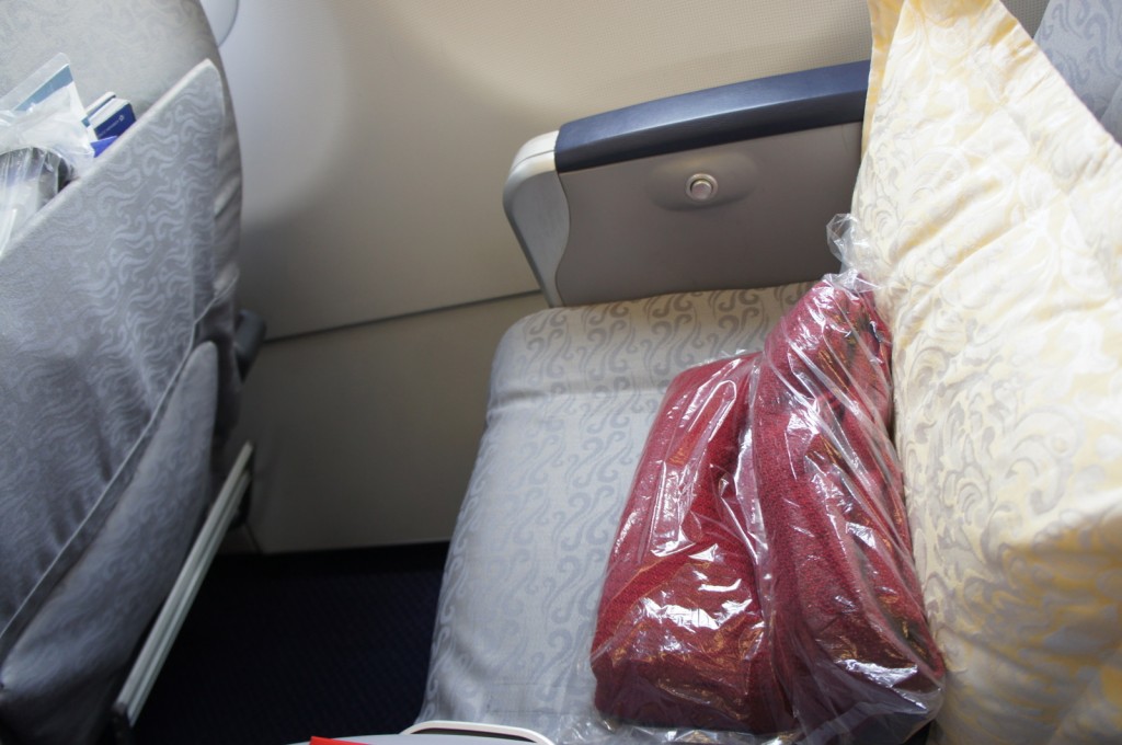 The image shows an airplane seat with a light gray patterned fabric. On the seat, there is a red blanket wrapped in plastic and a yellow pillow with a white floral pattern. The armrest is visible, and the back of the seat in front has a pocket containing some items.