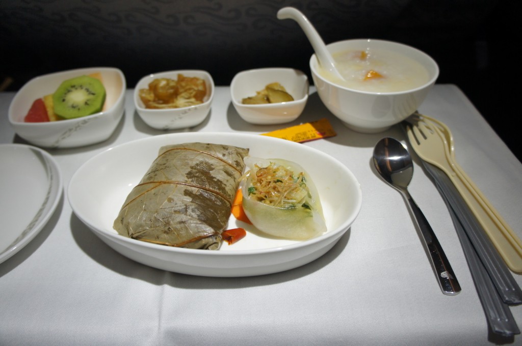 The image shows a meal served on an airplane. The meal includes a main dish wrapped in a leaf, a dumpling with vegetables, a bowl of congee (rice porridge) with a spoon, and several small side dishes including sliced fruit (kiwi and other fruits), pickled vegetables, and another small dish with a brownish food item. There are also utensils including a spoon, fork, and chopsticks placed on the right side of the tray. The meal is presented on a white tablecloth.