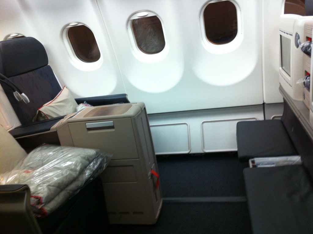 The image shows the interior of an airplane, specifically a business or first-class section. There are two large, cushioned seats with ample legroom and a console between them. The seats are equipped with pillows and blankets, which are wrapped in plastic. The area has three windows, and there is a small screen or monitor attached to the back of one of the seats. The overall setting appears clean and comfortable, designed for a premium travel experience.