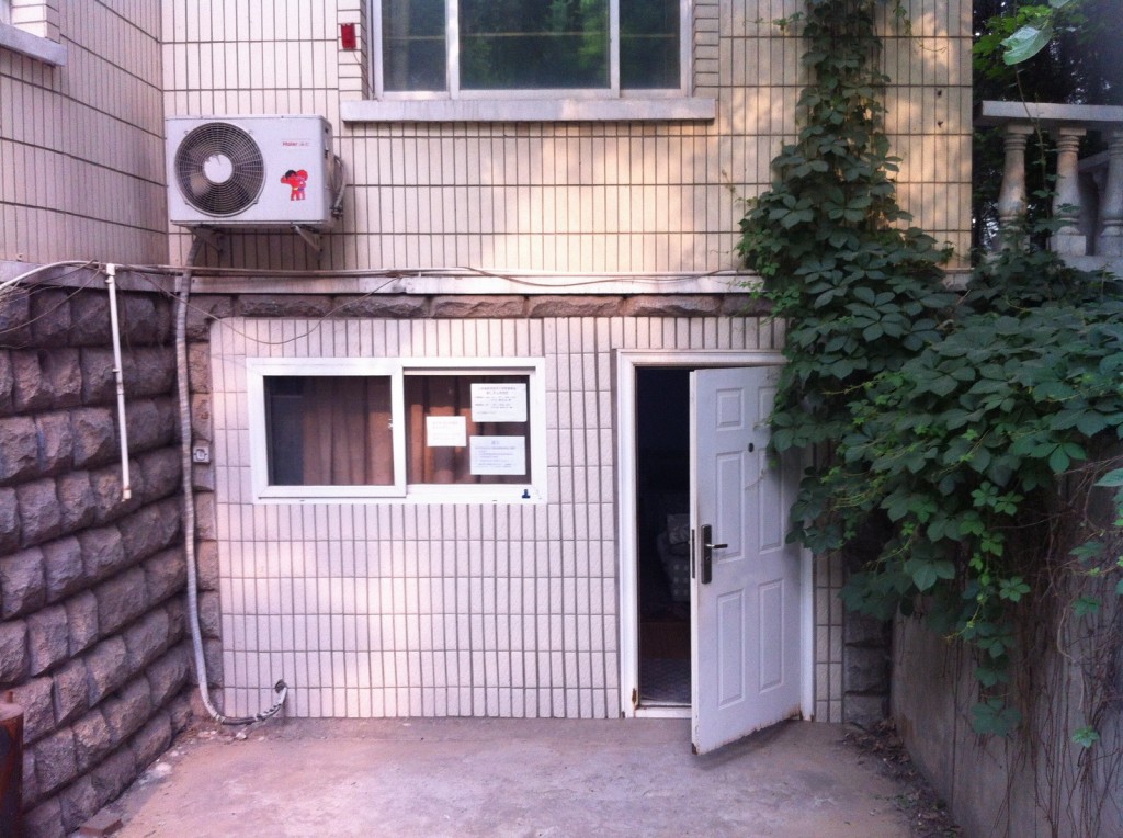 The image shows the exterior of a building with a tiled facade. There is a white door on the right side, which is slightly open, and a window on the left side with some papers posted on it. Above the window, there is an air conditioning unit mounted on the wall. The building is partially covered with green vines, especially on the right side near the door. The ground in front of the building is paved with concrete.