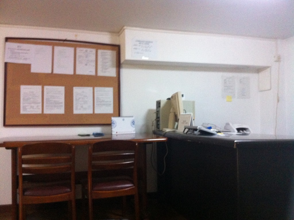 The image shows a small office space. On the left side, there is a wooden table with three chairs. Above the table, there is a large bulletin board with various papers and notices pinned to it. On the right side, there is a desk with an old computer monitor, a telephone, and some office supplies. The walls are white, and there are a few more papers and notices pinned to the wall above the desk. The overall setting appears to be a simple, functional office environment.
