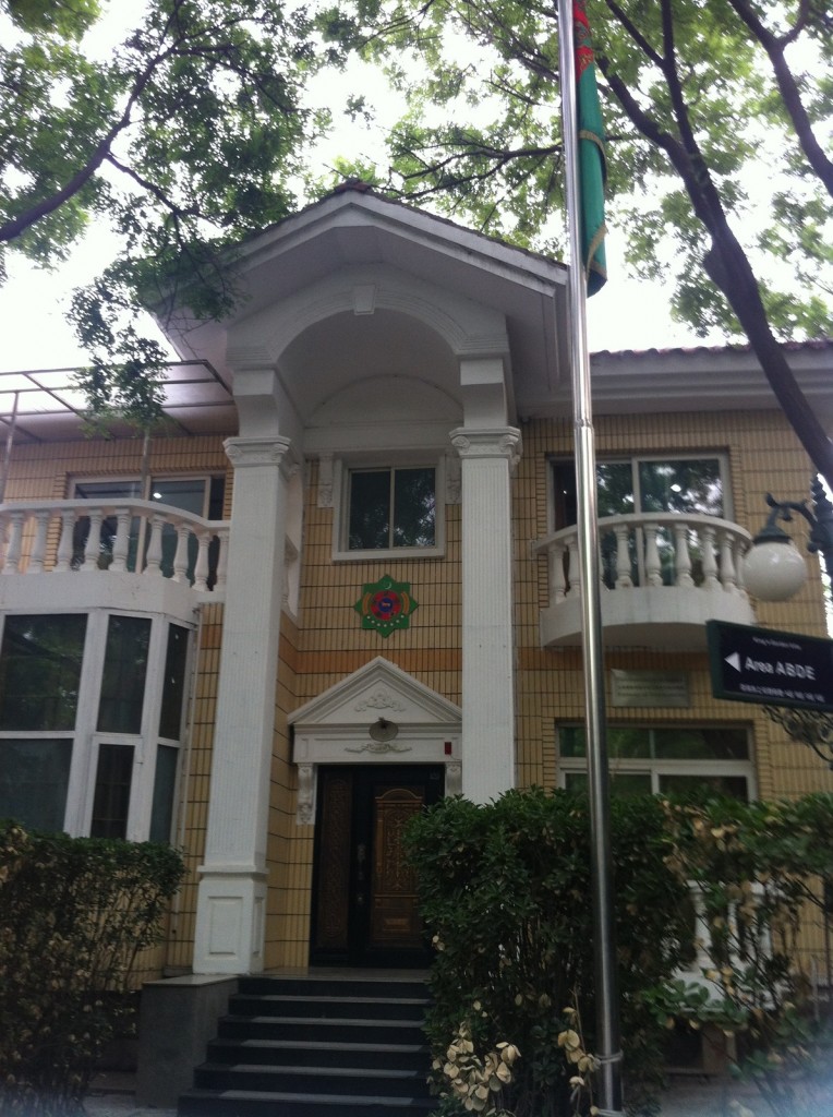 The image shows the entrance of a building with a classical architectural style. The building has a prominent portico with two large white columns supporting a triangular pediment. There is a decorative emblem above the door. The entrance door is dark and ornate, with steps leading up to it. On either side of the entrance, there are windows and small balconies with white railings. The building is surrounded by greenery, including trees and bushes. A flagpole with a green flag is visible in the foreground, and there is a sign on the right side of the image.