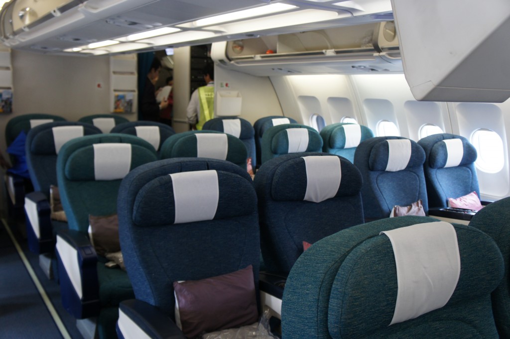 The image shows the interior of an airplane's business or first-class cabin. The seats are wide and upholstered in dark green or teal fabric, each with a white headrest cover. There are several rows of seats, and each seat has a pillow placed on it. The cabin is well-lit with overhead lighting, and there are windows along the side of the plane. In the background, a few people, possibly flight attendants or passengers, are standing and interacting near the entrance to another section of the plane.