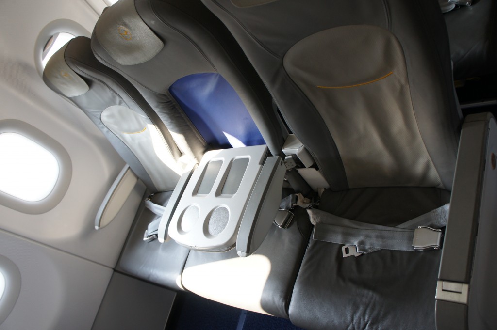 The image shows a row of three airplane seats. The seats are upholstered in dark gray leather with light gray headrests and seatbacks. The middle seat has its tray table down, which has two cup holders and a small rectangular indentation. The seats are equipped with seat belts, and the window on the left side of the image allows natural light to enter the cabin. The overall setting appears to be the interior of an airplane.