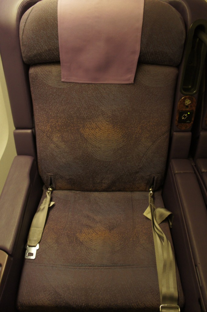 The image shows a close-up of a single airplane seat. The seat is upholstered in a dark fabric with a subtle pattern and has a light purple headrest cover. The seatbelt is visible, with the straps lying on the seat. The armrests are also visible, with one side showing a control panel.
