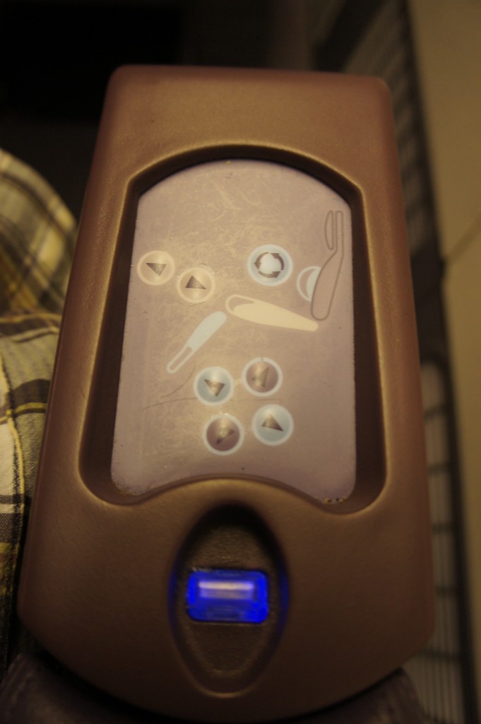 The image shows a close-up of a control panel, likely for adjusting a seat. The panel has several buttons with icons indicating different seat adjustments, such as reclining the backrest, raising or lowering the leg rest, and adjusting the seat position. The buttons are arranged in a circular pattern, and there is a blue light indicator at the bottom of the panel. The control panel is encased in a brown plastic housing.