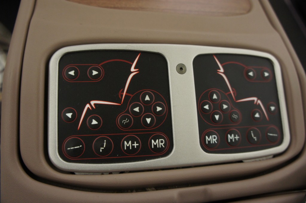 The image shows a close-up of a control panel for adjusting a car seat. The panel has two sections, each with various buttons. The buttons include arrows for adjusting the seat position, symbols for lumbar support, and other seat functions. The panel is set in a beige-colored car interior.