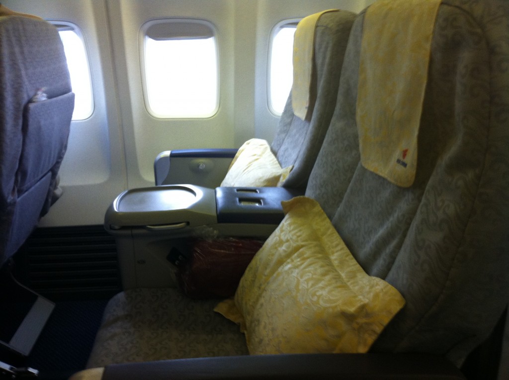 The image shows a row of airplane seats in the economy or business class section. The seats are upholstered in a patterned fabric and have light yellow headrest covers and pillows. There are two windows visible in the background, and the seats have armrests with built-in tray tables. The overall setting appears to be clean and well-maintained.