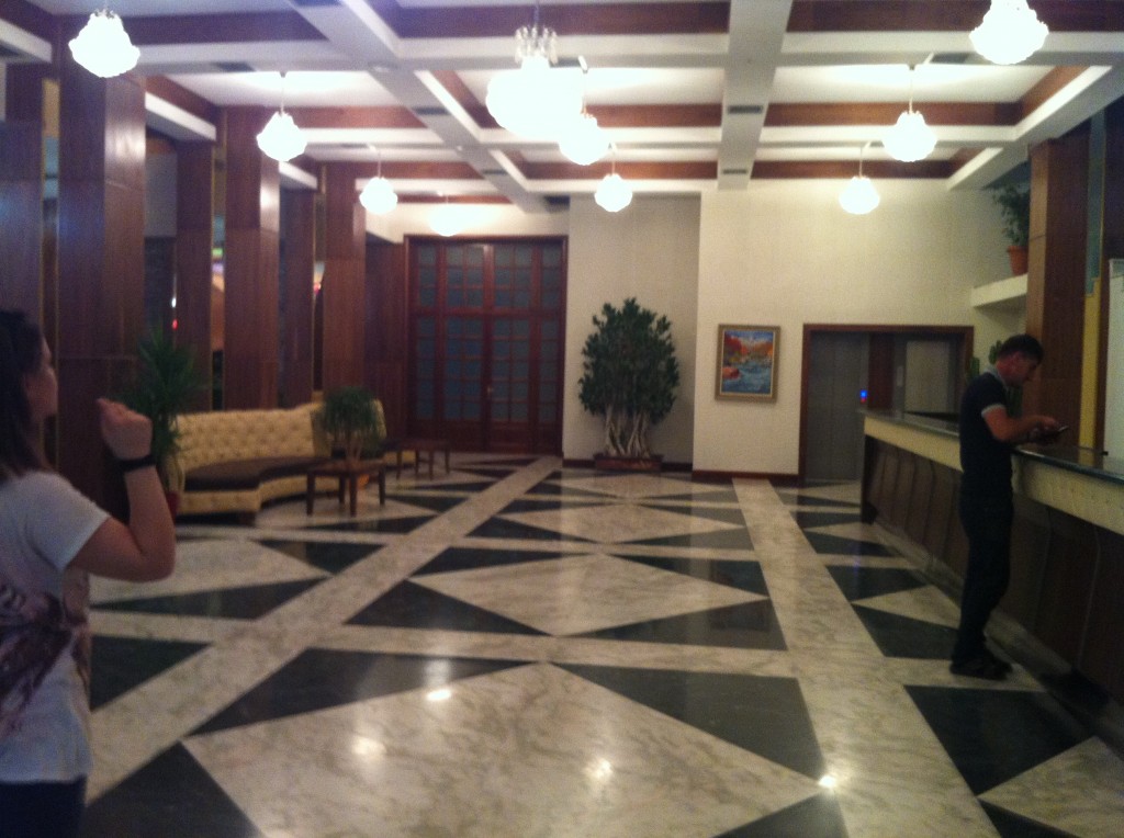 The image shows the interior of a spacious, well-lit hotel lobby. The floor has a geometric black and white pattern. There are several chandeliers hanging from the ceiling, providing ample lighting. On the left side, there is a woman standing and looking towards the lobby, while on the right side, a man is standing at the reception desk, possibly checking in or out. In the background, there is a seating area with a beige sofa and a couple of chairs, along with some potted plants. A large plant is also visible against the back wall, which has a framed painting and a set of double doors.