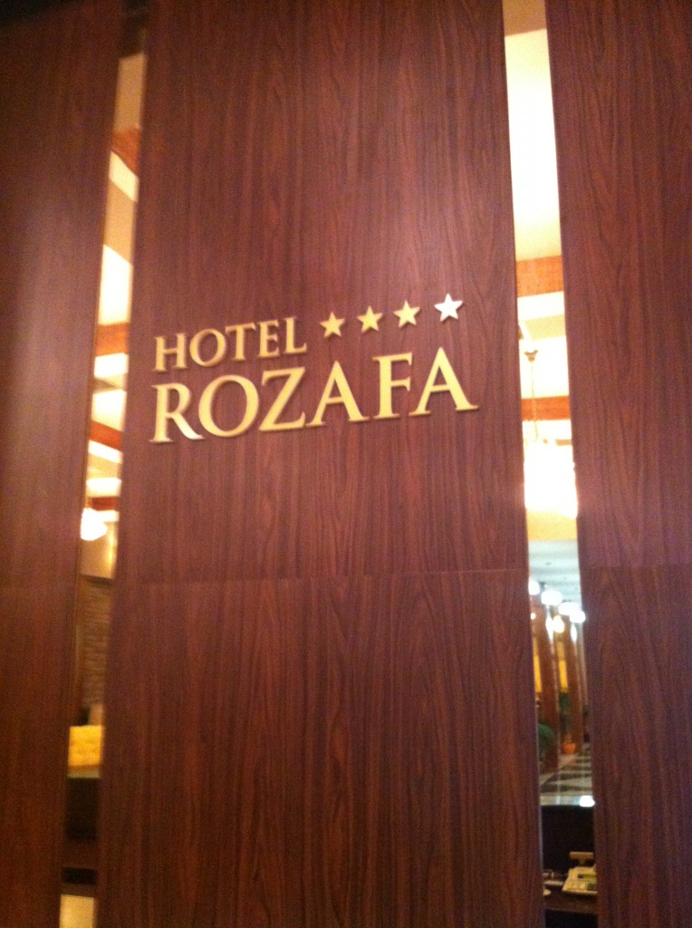 The image shows a wooden wall with the text "HOTEL ROZAFA" in gold letters. Above the text, there are four gold stars, indicating a four-star rating for the hotel. The background appears to be part of the hotel's interior, with some lighting and decor visible.