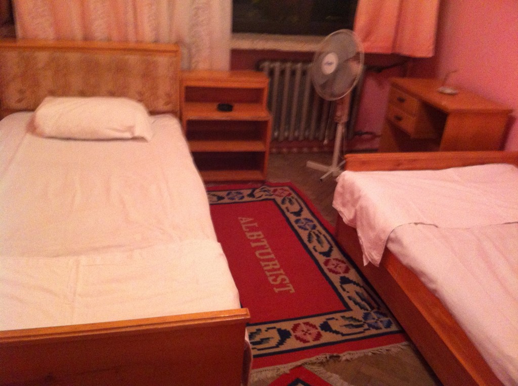 The image shows a small bedroom with two single beds. The beds are positioned parallel to each other with a wooden nightstand in between. Both beds have white sheets and pillows. There is a red and blue patterned rug on the floor with the word "ALBATURIST" visible. A standing fan is placed near the window, which has pink curtains. There is also a wooden dresser with drawers on the right side of the room. The walls are painted pink.