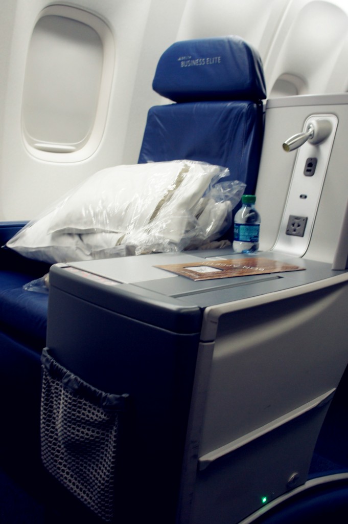 The image shows a business class seat on an airplane. The seat is upholstered in dark blue leather and has a headrest with the words "BUSINESS ELITE" printed on it. There is a large pillow wrapped in plastic placed on the seat. Next to the seat, there is a small table with a bottle of water and a menu or pamphlet on it. The area also features a power outlet and a small storage pocket. The window of the airplane is visible in the background.