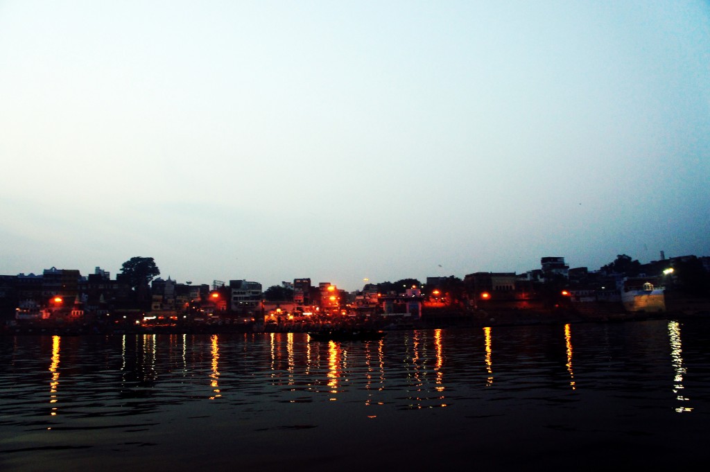 Alt text: A serene evening view of a riverside town with buildings illuminated by streetlights. The lights reflect off the calm water, creating a picturesque scene. The sky is clear, transitioning from light to dark as night approaches.