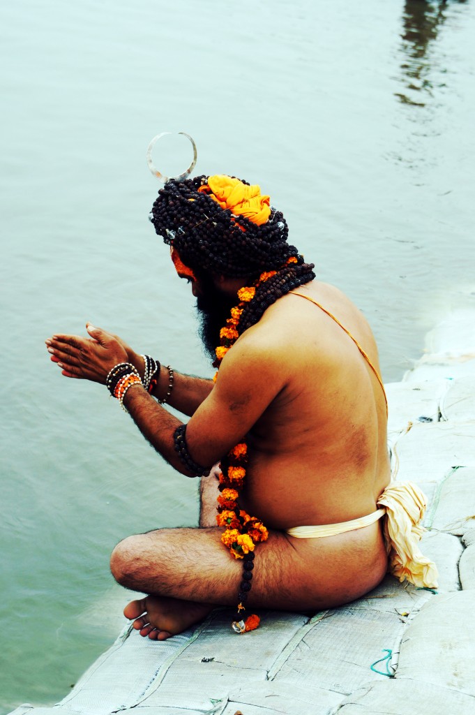 A person with a beaded headdress and garlands of marigold flowers around their neck and wrists is sitting by the water, possibly engaged in a ritual or prayer. The individual is wearing minimal clothing and appears to be in a meditative or prayerful pose with hands clasped together. The background shows a body of water.