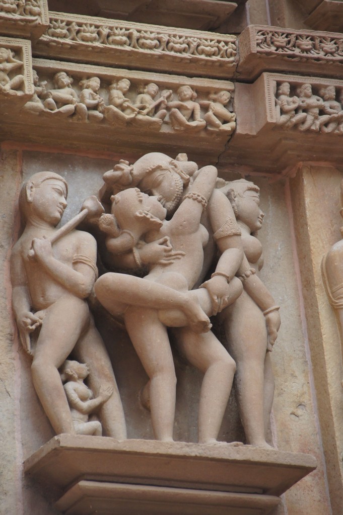 The image shows an intricate stone carving on a temple wall, depicting three human figures in an intimate and complex pose. The figures are intricately detailed, with one figure in the center being held by the other two. Above the main figures, there is a frieze with smaller carvings of seated and standing human figures engaged in various activities. The carving is part of a larger architectural structure, likely of historical and cultural significance.