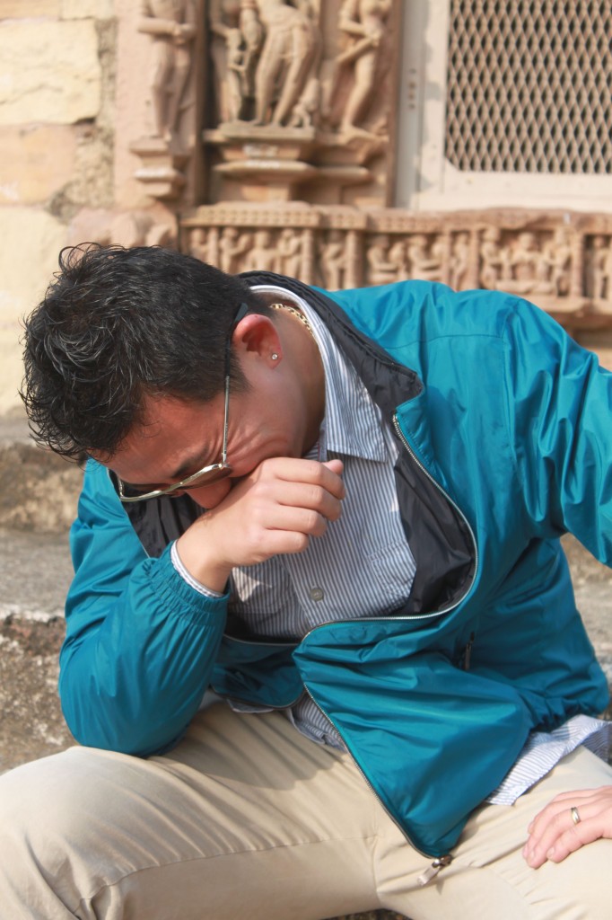 A person wearing a blue jacket, striped shirt, and beige pants is sitting down with their head bowed and hand covering their face, appearing to be in distress or deep thought. The background features intricate stone carvings and a mesh window.