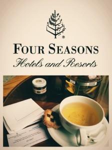 The image shows a promotional picture for Four Seasons Hotels and Resorts. The top part of the image features the Four Seasons logo and the text "Four Seasons Hotels and Resorts." Below the text, there is a photograph of a cup of tea on a saucer with a cookie on the side. Next to the cup, there is a small jar of jam and some papers, possibly a brochure or a menu. The setting appears to be a cozy and elegant environment, likely within a hotel or resort.