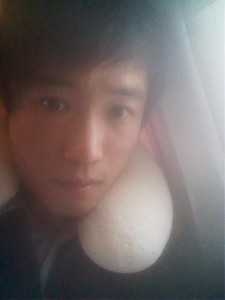 A person is taking a selfie while sitting next to a window. They are wearing a neck pillow, suggesting they might be on a plane or another form of transportation. The lighting is soft and slightly dim.